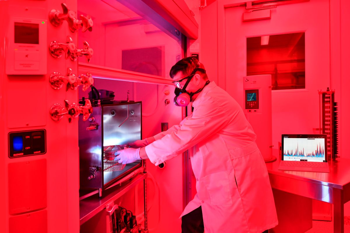 This is a lab lit with red lighting. In the middle, a person in a white lab coat waring a protective mask reaches into a counter space