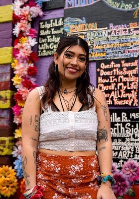 Photograph of scholarship recipient Mia Sebastian. She is pictured in front of a colorful graffitied wall decorated with artificial flowers. She is wearing a white lace cropped shirt and an orange skirt. She has long black hair and small tattoos.