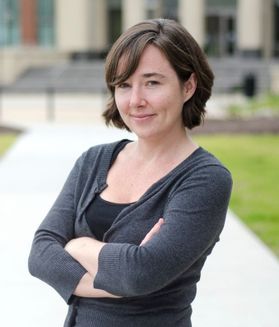 Headshot of WVU professor Ember Morrissey. She is pictured standing outside with her arms crossed. She is wearing a gray sweater and a black shirt. She has short brown hair just above her shoulders and is slightly smiling. 