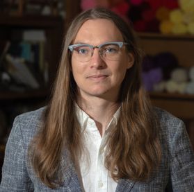 Headshot of WVU research assistant Jordan Masters. They are pictured inside wearing a gray tweed coat over a white, button-up dress shirt. They have long, light brown hair and are wearing gray framed glasses. 