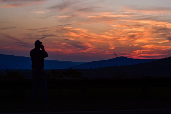 A person is shown in silhouette while taking a photo of a sunset over mountains