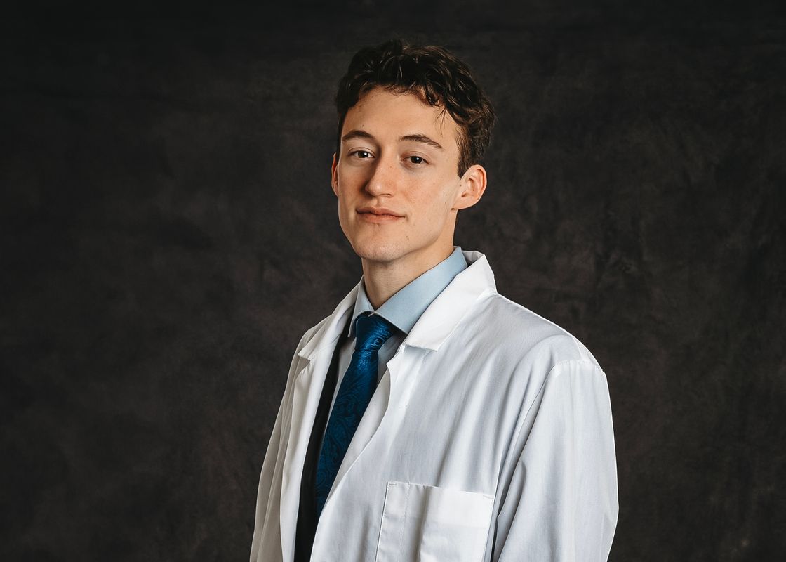 Photograph of Easton Cahill, an undergraduate researcher at WVU. He is pictured against a black background wearing a white lab coat over a light blue shirt and bright blue tie. He has curly brown hair. 