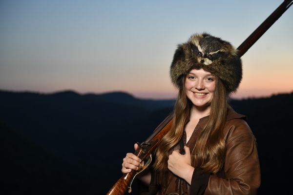 A woman with brown hair wearing a brown leather outfit with a furry coon hat. She is standing in front of a sunset. The sky is pastel blue, pink, and orange.