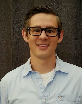 Photo of man with dark hair and black glasses in blue button down shirt standing in front of dark brown wall