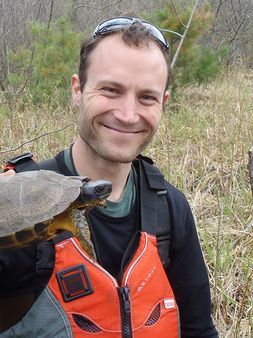 A person wearing a life jacket looks at the camera while holding a turtle up to it. The person is outside and smiling with glasses on their head.