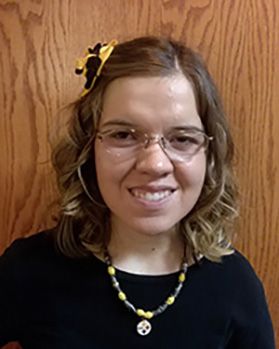 Headshot of WVU employee Mary Veselicky. She is pictured in front of a wooden wall and is wearing a black shirt with a Pittsburgh Steelers necklace and matching black and yellow bow in her hair. She has shoulder length light brown hair and wears glasses. 