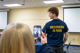 A male student poses wearing a T-shirt that says "Recovery is rad" while a female student with blonde hair takes his photo. 