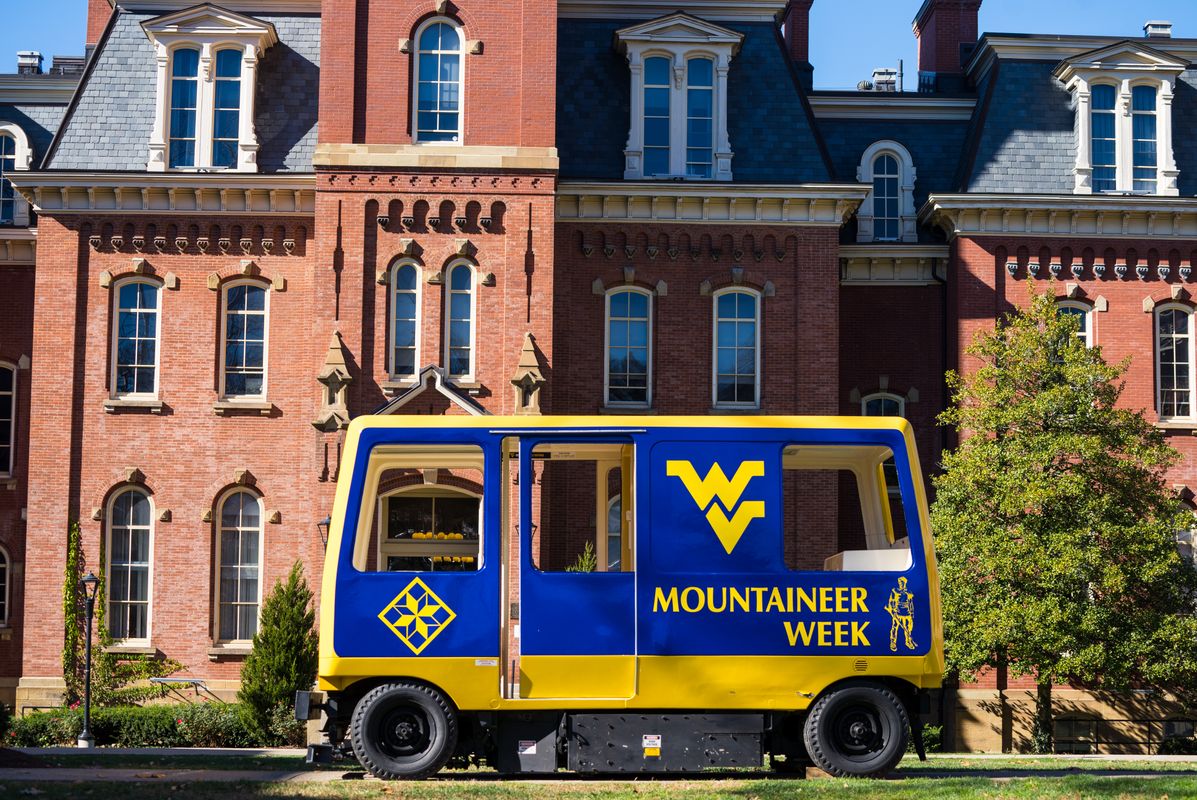 A West Virginia University branded PRT cart is parked in Woodburn Circle with the iconic Woodburn Hall in the background. The PRT cart says Mountaineer Week on it. 