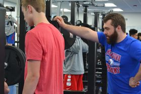 A man in a blue t-shirt helps students with weight training