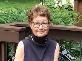 WVU College of Law almuna Ellen Archibald is pictured here seated outside, wearing a sleeveless blouse, circle-rimmed glasses, and short, curly brown hair.