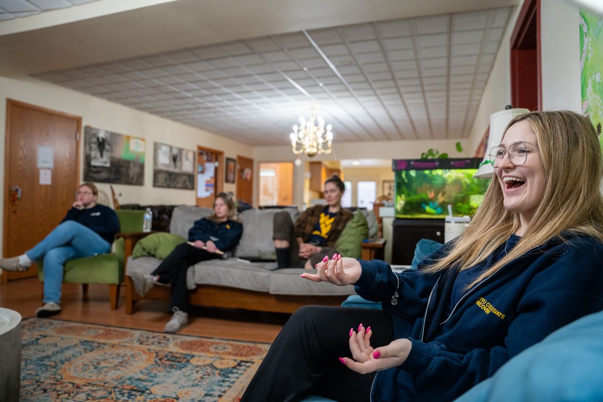 A group of students sit together in a comfortable room with couches and chairs and a illuminated fish tank. A girl with blonde hair is laughing and gesturing with her hands. 