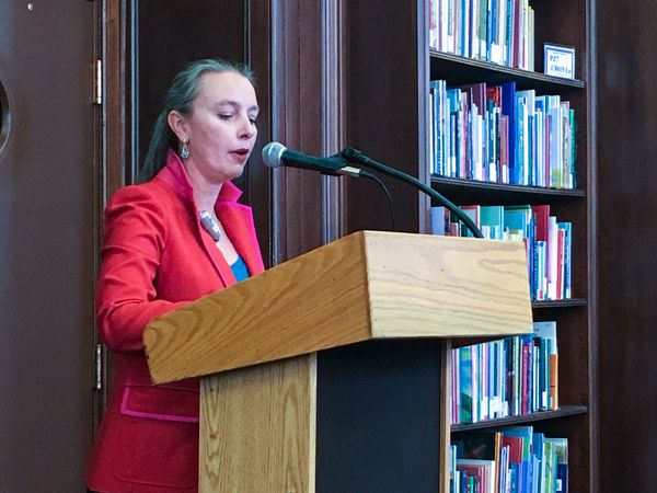 woman stands in red jacket speaking at microphone/podium with books in the background