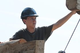 boy in hard hat raises arm to lift piece of wood