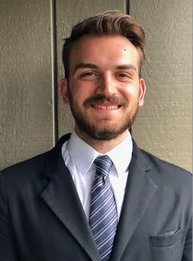 Headshot of WVU graduate student Evan Cramer. He is pictured against a beige background and is wearing a gray suit over a white dress shirt and blue striped tie. He has short brown hair and a brown beard. 