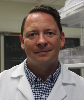 Headshot of WVU researcher Aaron Robart. He is pictured inside of a lab space wearing a white lab coat over a dark blue gingham shirt. He has short brown hair. 