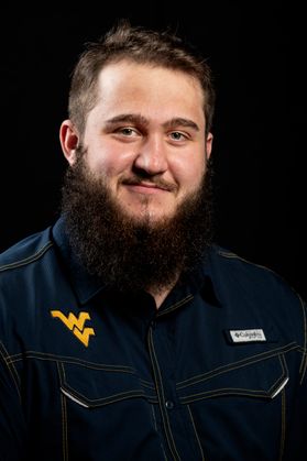 Photo of a young man with dark hair and beard wearing a dark shirt with a flying WV against a dark background