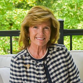 Headshot of WVU administrator Cindi Roth. She is pictured seated on a white couch with green trees and a black metal fence behind her. She is wearing a black and white checkered jacket over a black shirt. She has shoulder length strawberry blonde hair 