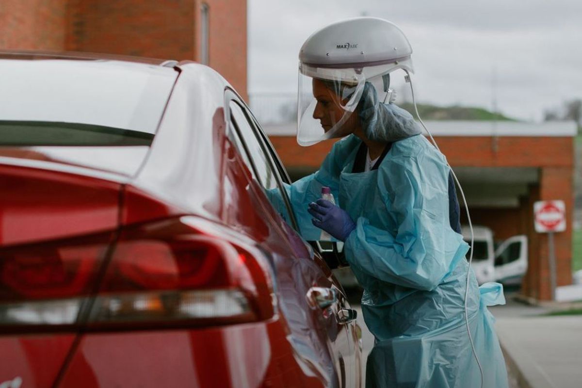 Woman in green medical protective gear administers COVID-19 test to a patient in a red car