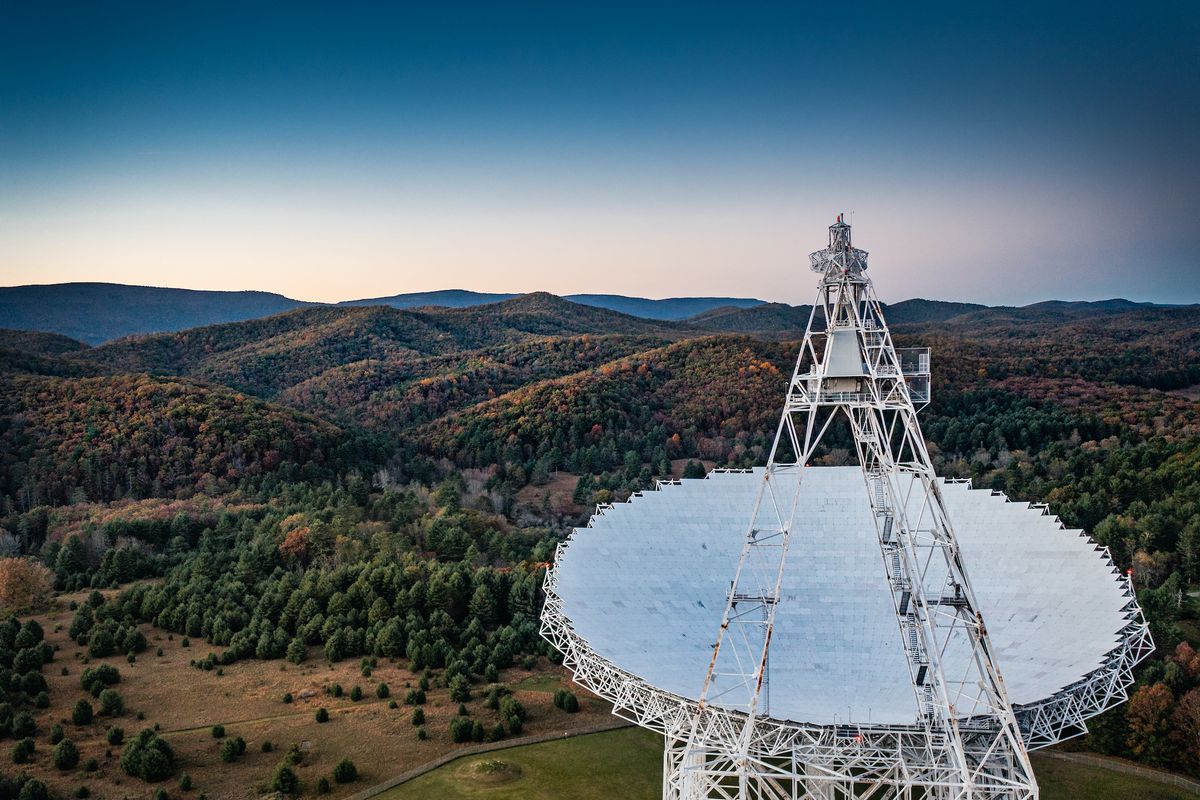An aerial image of the Green Bank Telescope in rural West Virginia. A mountain range and thick green forest can be seen in the distance behind the telescope. 