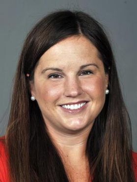 This is portrait of April Messerly who has long dark hair and is wearing a red shirt and white pearl earrings while sitting in front of a gray backdrop.