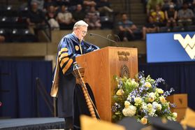 Gordon Gee speaking at a podium for graduation. He is wearing a blue gown and has grey hair with glasses.