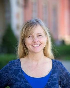 Headshot of WVU professor Dana Coester. Dana is pictured outside with red brick buildings in the background. She is wearing a royal blue top and has long, blonde hair. She is smiling in the photo. 