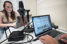 An image taken in a computer lab of students working on laptops. A microphone sits on the table between them. You can see one of the laptop screens measuring sound. 