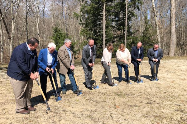 Seven people stand with shovels in the dirt to break ground in a field with trees in the background