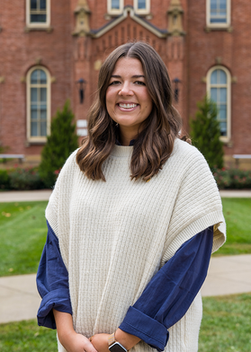 A person with long dark hair stands outside Woodburn Hall wearing a long-sleeved blue shirt with a white sweater over top of it.