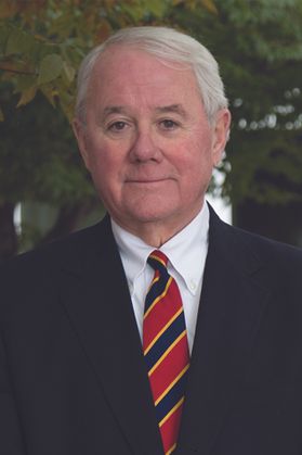 Headshot of WVU professor James Wood. He is standing outside in front of green trees. He is wearing a dark suit jacket over a white dress shirt with a red, blue and gold striped tie. He has short gray hair. 