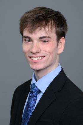 Headshot of WVU Bucklew Scholar Nathaniel Lange. He is pictured against a gray background wearing a dark colored jacket over a light blue dress shirt with a blue patterned tie. He has short, dark hair. 