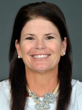 Headshot of WVU women's soccer coach Nikki Izzo-Brown. She is pictured against a gray background and is wearing a white blouse with a large necklace. She has very long brown hair and is wearing pearl earrings. 
