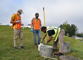 Men stand on grassy hillside working around a hole, concrete cylinder in foreground