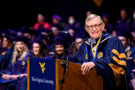 WVU President Gordon Gee stands behind a microphone at a wooden podium. He is wearing gold and blue academic regalia and smiling.