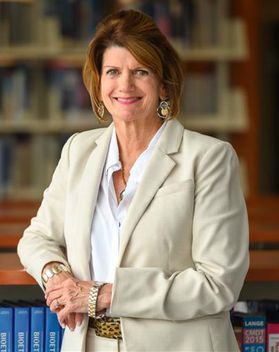 Headshot of retiring WVU administrator Laura Gibson. She is pictured inside wearing a cream colored pant suit over a white dress shirt. She has short, auburn hair and is wearing dangling earrings. 