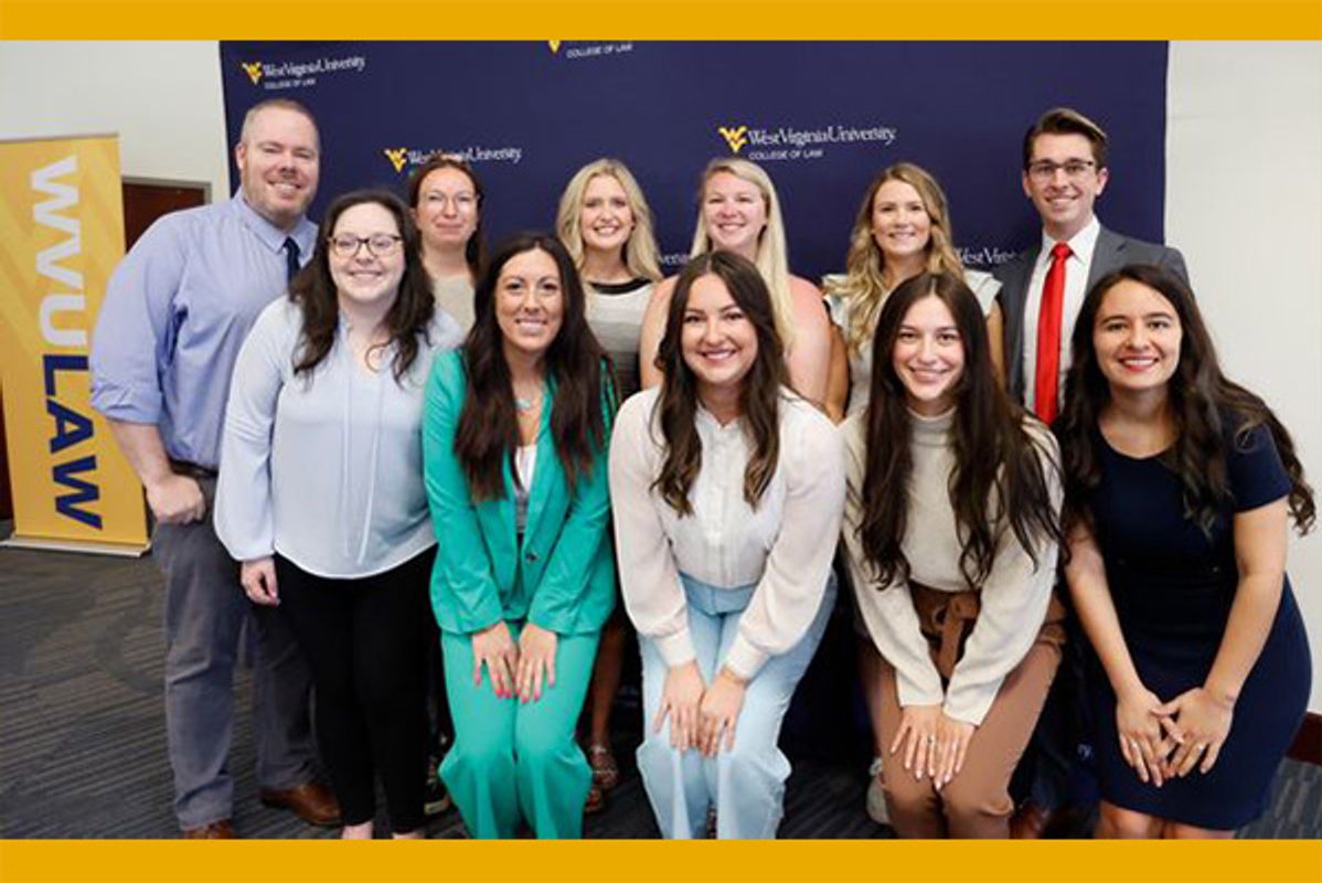A group of WVU College of Law graduates posing for a group picture. Their attire is business casual as they smile in front of a navy WVU backdrop.