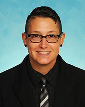 Headshot of WVU researcher Dr. Nova Szoka. Dr. Szoka is pictured against a light blue background and is wearing a dark coat with a gray striped tie, short dark hair, and glasses. 