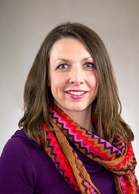 Headshot of WVU professor Kristin McCartney. She is pictured in front of a beige background wearing a purple top with a coordinating colorful scarf around her neck. She has shoulder length brown hair. 