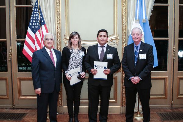 Three men and one woman stand wearing suits in front of white and gold bordered window with the American and Argentinian flags behind them