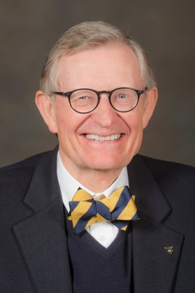 Headshot of WVU President Gordon Gee. He is pictured against a dark beige background. He is wearing a navy blue suit with a navy blue sweater vest and a blue and gold bowtie. 