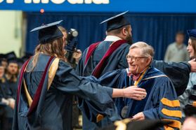 President Gordon Gee, dressed in gold and blue commencement regalia, opens his arms to hug a graduate who is wearing a blue graduate gown and blue mortarboard.