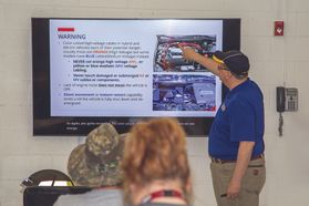 A man in a blue shirt stands next to a video screen and instructs a group of people.