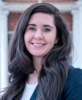 Headshot of WVU administrator Erin Newmeyer. She is pictured outside wearing a navy blue jacket over a white blouse. Her long wavy dark hair flows over her shoulders. 