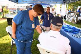A nurse in blue scrubs with short auburn hair works with a male patient seated in a chair under an outdoor tent during the Helping Harrison health campaign. 