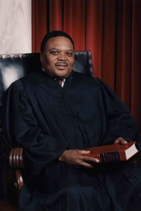 man in black judge's robe sits in black chair holding book
