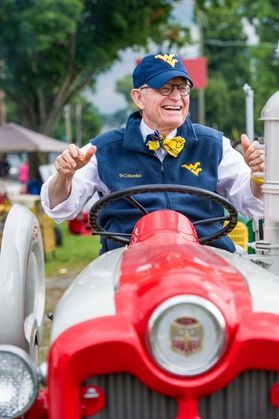 An older man with gray hair and glasses operating a red and silver tractor. He's wearing a white button up shirt with a blue vest and a yellow and blue bow tie.