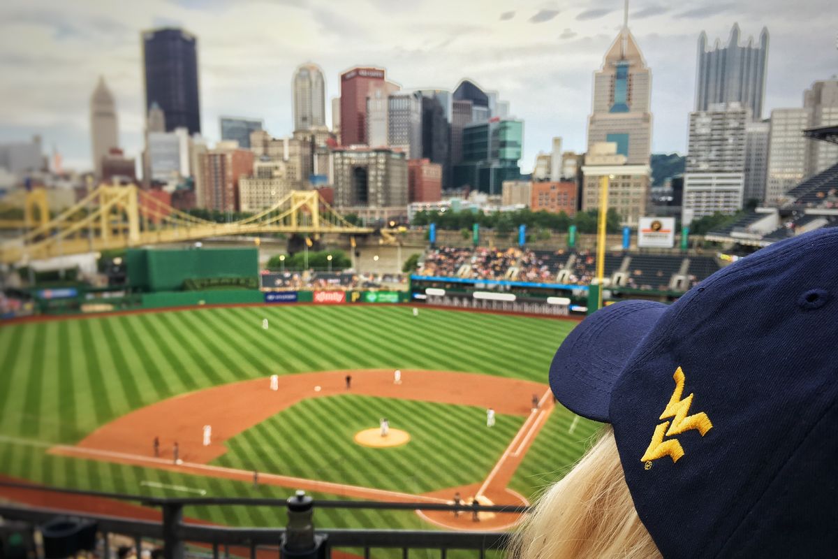 Photo of a baseball diamond with a city-scape in the background, dark blue hat with flying WV in the foreground