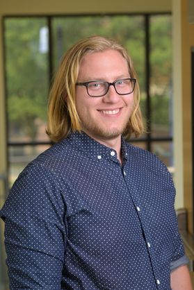 WVU student Andrew Kaiser. He is pictured in front of a window wearing a navy blue button-up shirt with tiny white dots. He has shoulder length blonde hair and wears black square-framed glasses. 