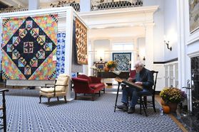 A quilt show held on the WVU Campus. Colorful quilts in many colors and patterns are hung on the walls of an ornate room. An older man sits in a chair in the corner of the room playing an instrument. 