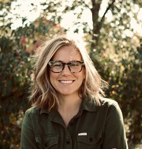 Headshot of WVU employee Morgan Haas. She is pictured outside with full green trees behind her. She is wearing an olive green button up shirt and has shoulder length blonde hair. She also wears tortoise framed glasses. 